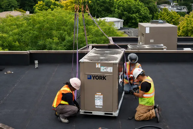 Three crew members in hard hats and high-visibility clothing stand beside a rooftop HVAC unit prepared for a crane lift.