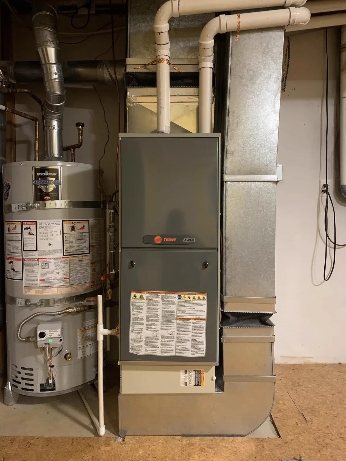 A gas furnace installed beside a water heater in a utility space, with the flue and gas piping run overhead.
