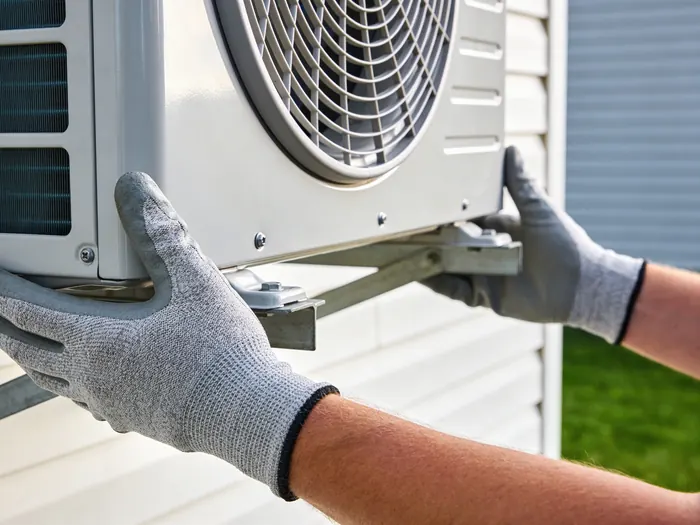 Two gloved hands supporting an outdoor heat pump unit onto a wall bracket against pale siding.