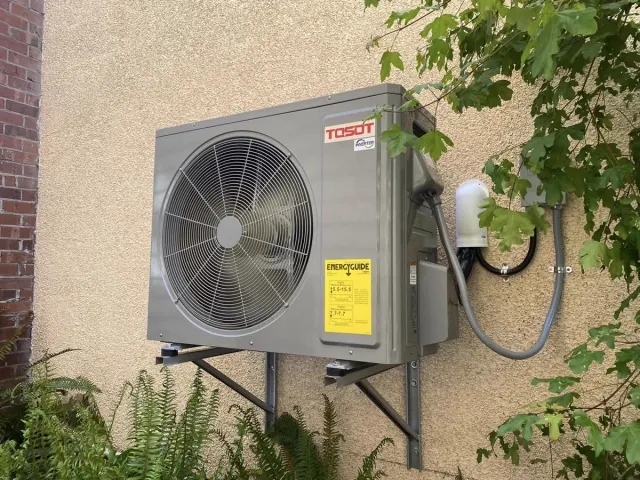 An outdoor heat pump unit carried on steel wall brackets against a stucco wall, with the refrigerant lines and an electrical disconnect beside it.