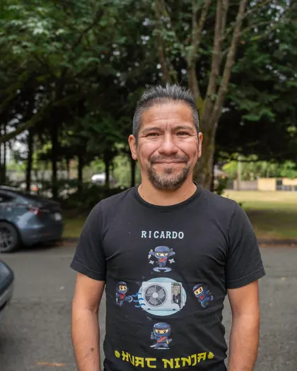 A man standing outdoors in a parking area, facing camera, in a dark Cool Guys crew t-shirt printed with the first name Ricardo. Trees behind; the company van is partly visible at the right edge.