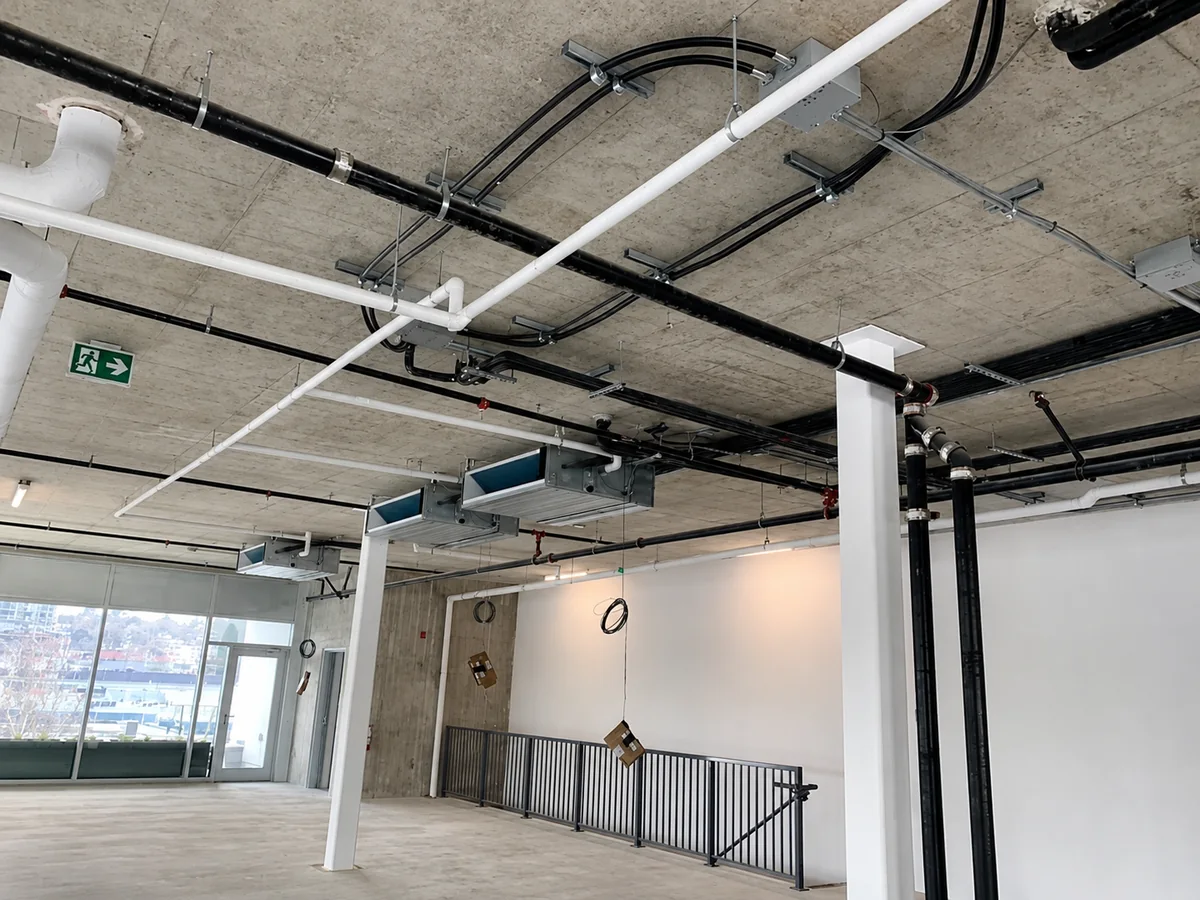 An unfinished commercial interior with exposed concrete, suspended ventilation equipment, ductwork, piping, and electrical runs overhead.