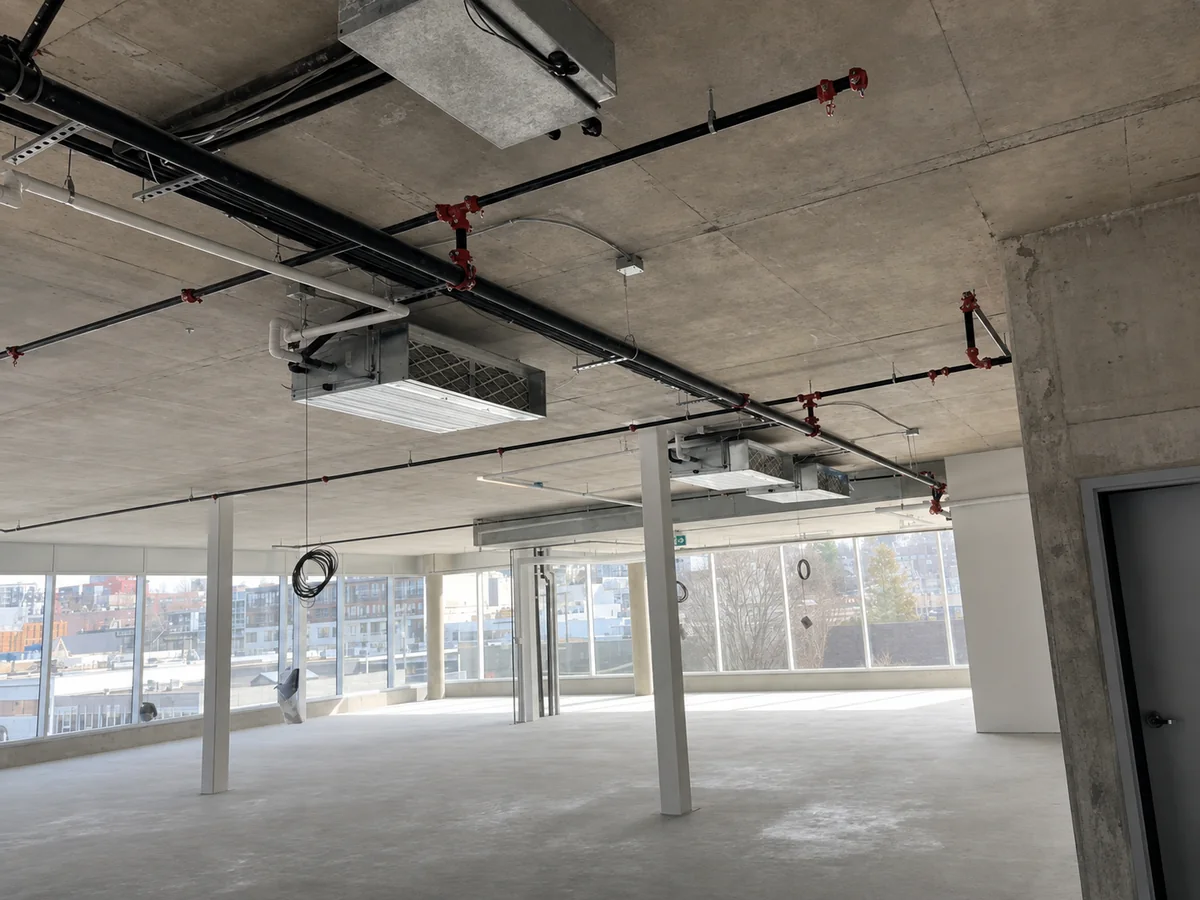 A wide open commercial floor with ventilation equipment, ductwork, piping, and electrical services suspended below a concrete ceiling.