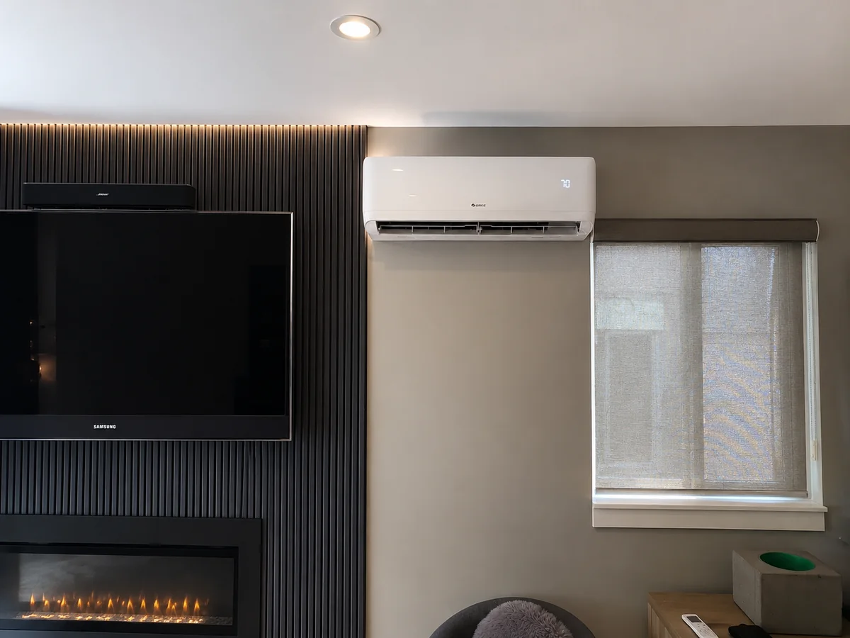 A wall-mounted indoor ductless unit above a window beside a dark fireplace and television feature wall in a furnished living room.